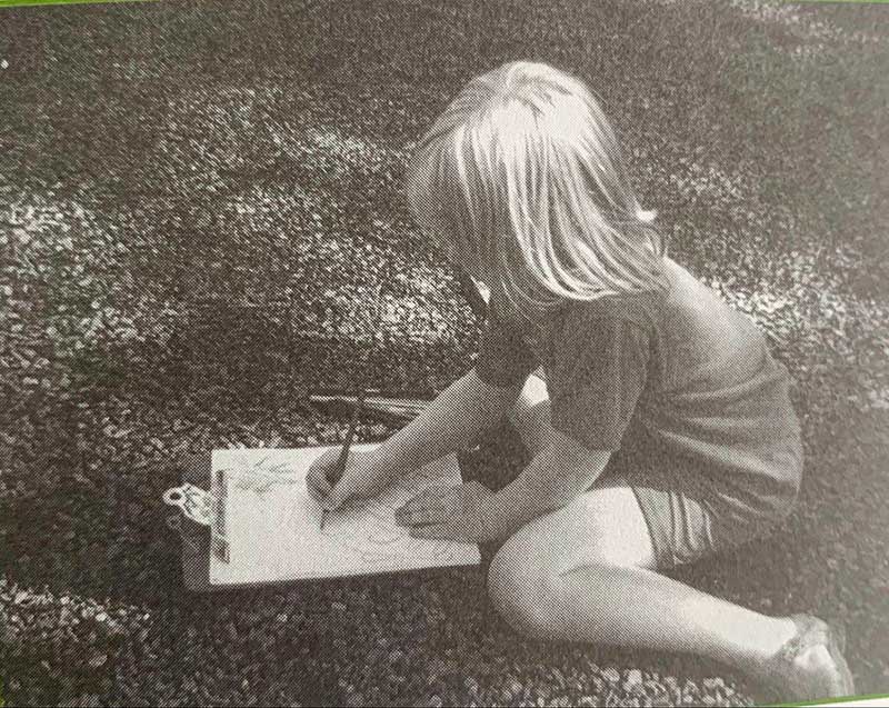 Black-and-white photo of a child drawing on a clipboard while sitting on the grass