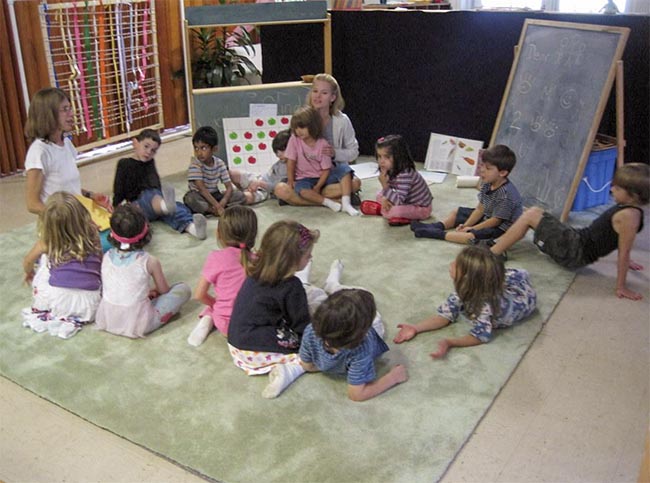 A group of young school-aged children sit in a circle sharing while their teacher looks on and guides the group.