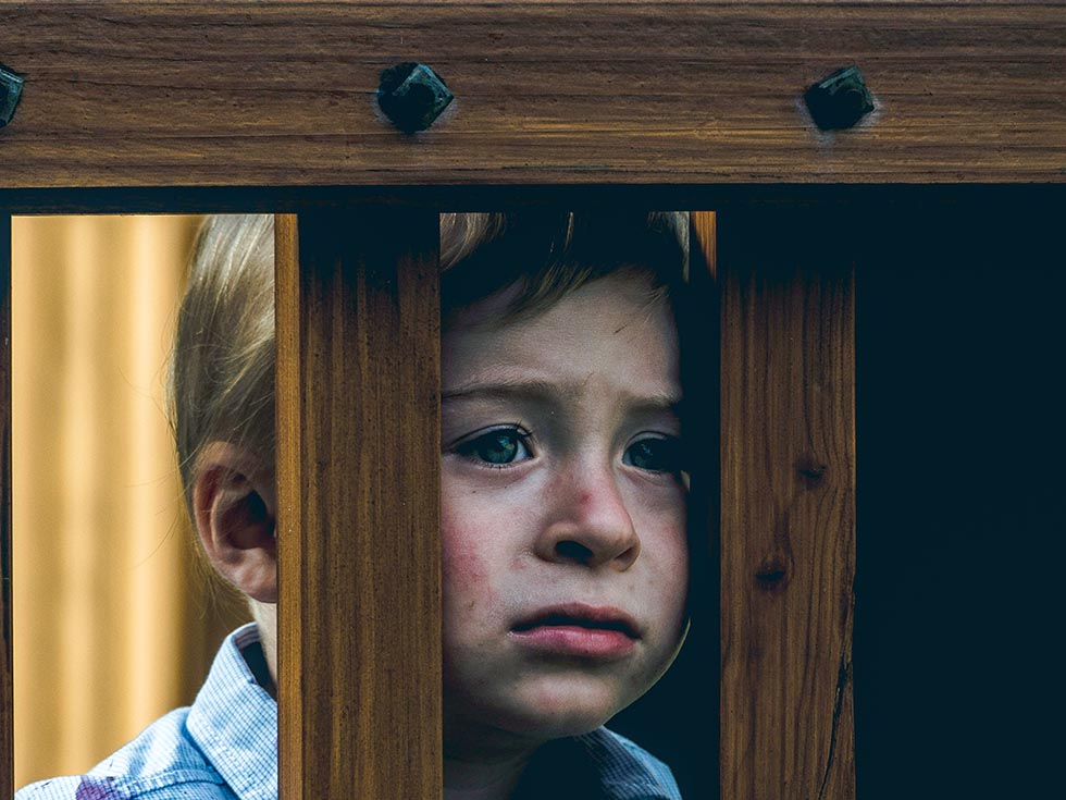 A distressed-looking young boy peers through 2 slats of a fence.