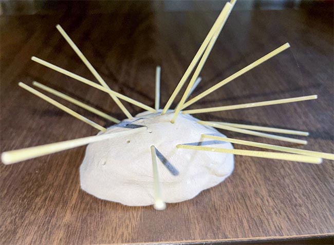 A mound of clay sits on a schoolroom table with long sticks of spaghetti sticking out at all angles.