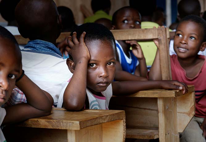 A group of young African school boys sit in wooden benches. One boy turns around to face the camera leaning his head in his hand with his elbow propped up on the bench.