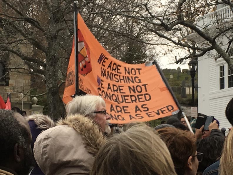 Parade of protestors holding an orange sign reading, "We are not conquered; we are not vanishing"