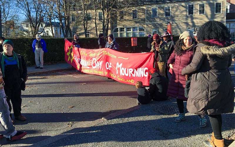 Group of marchers hold a long red banner reading: International Day of Mourning. It's cold outside, so people are bundled up in jackets and scarves.