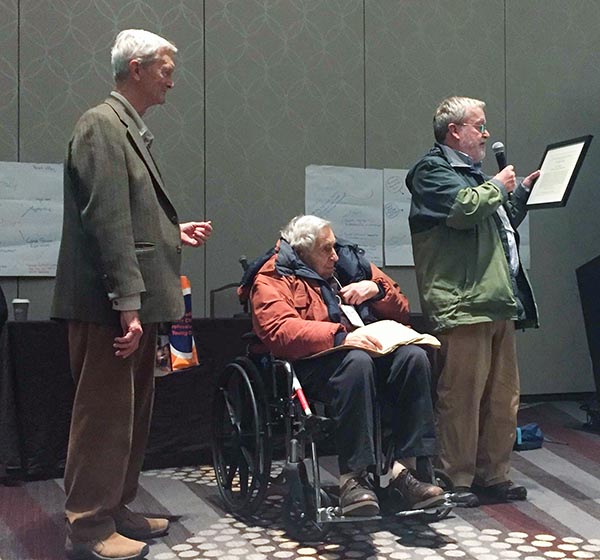 A senior man in a wheelchair receives an award. One man stands by listening and one man speaks into a microphone.