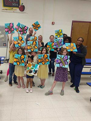 A group of adults and children in a classroom. they all hold up small canvases with paintings of a butterfly