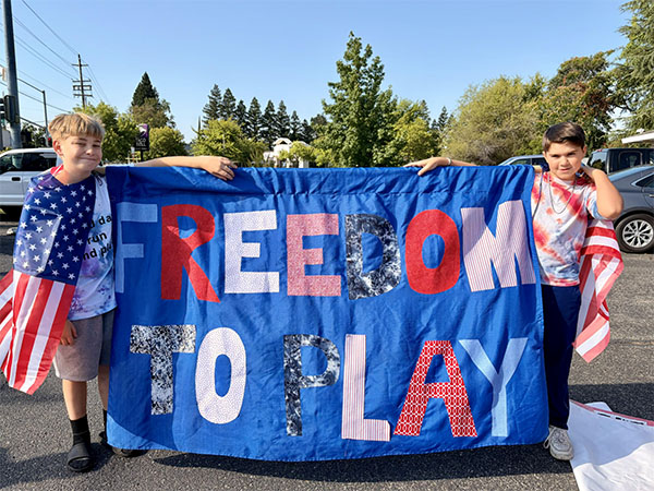 Two school-aged boys hold a parade sign that reads 'Freedom to Play.' Both boys were a US flags as capes.