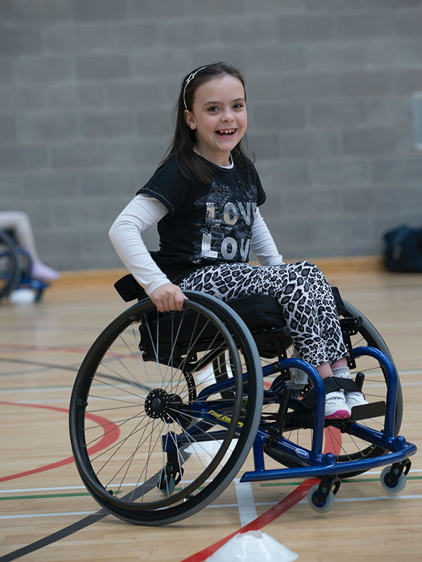 An elementary-aged girl in a wheelchair plays basketball oon an indoor court.