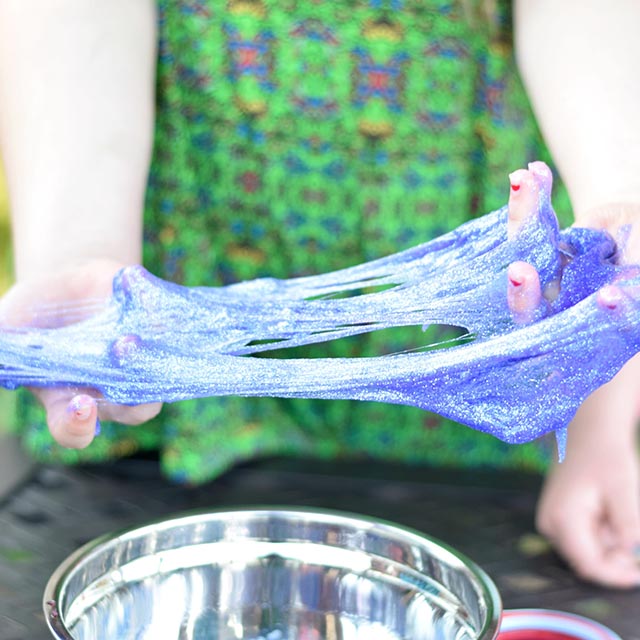 A young girl stretches glittery purple slime between her hands.