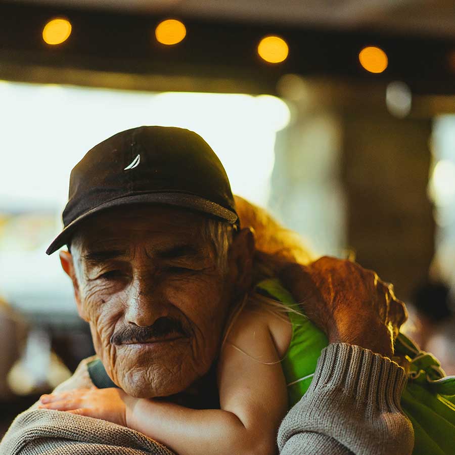 A grandfather in a baseball cap hugs a young girl to him. Her head is behind his and she drapes over his shoulder.