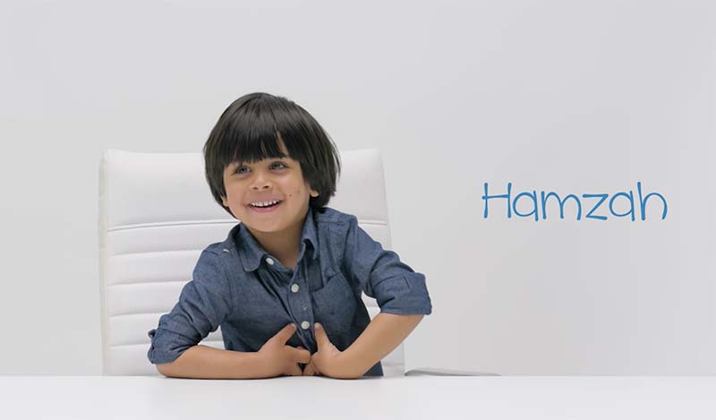 A young Middle-Eastern boy sits at a white table in a white chair wearing a long-sleeved blue shirt. The boy smiles at the camera.