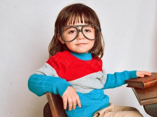 Young child in a striped sweatshirt with an adult's eyeglasses on. The child sits at a desk and has a hand on top of a book on the desk.