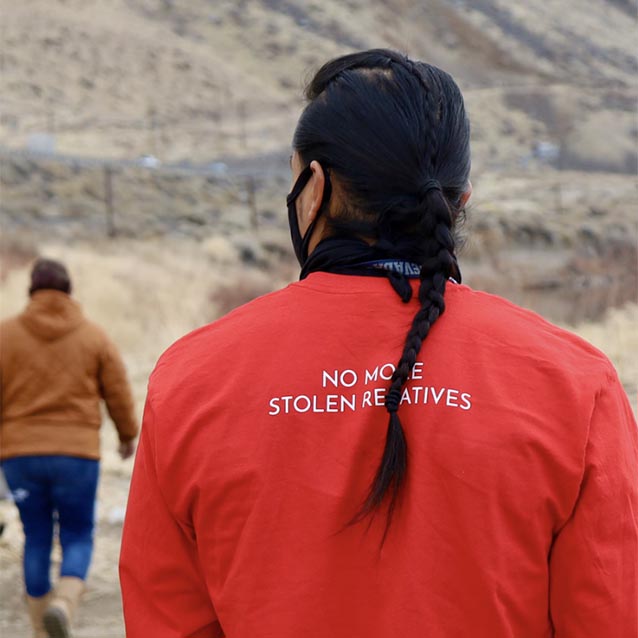 The back of an indigenous man in a red coat which says no more stolen lands.