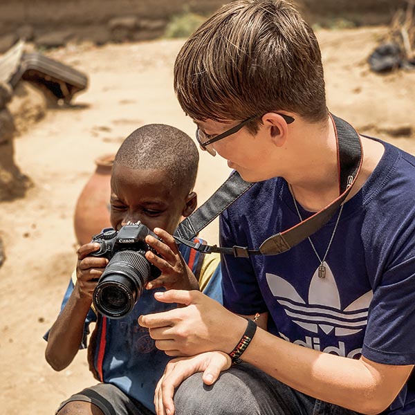 A woman bends down to let a young African boy look through her camera