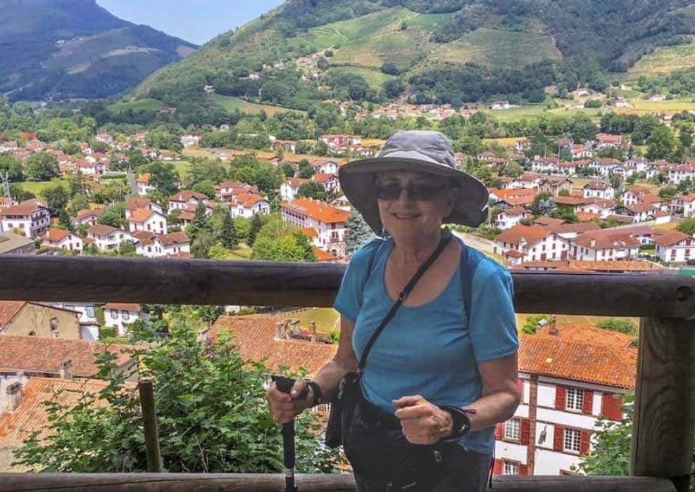 Older Caucasian woman smiles at the camera. She wears hiking gear and has a walking stick. Behind her is a quaint town with terracotta roofs and green hills.