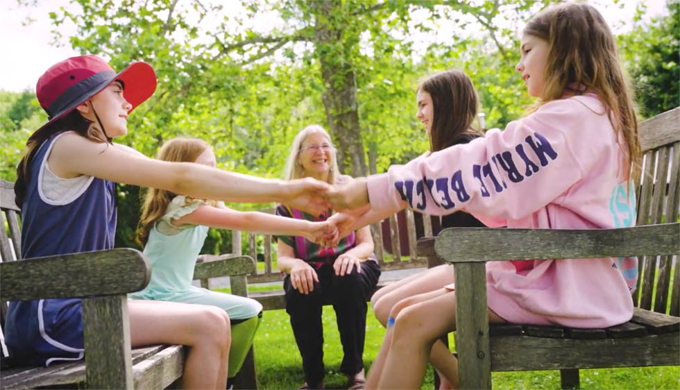 An older woman leading 4 girls in camp songs. The girls sit on benches across from each other and hold hands.