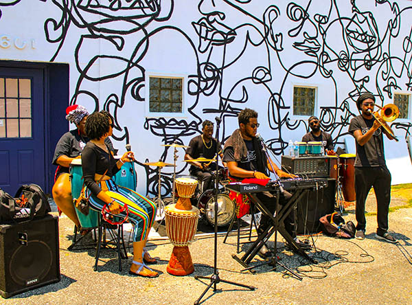 A 6-piece band plays at an outside festival in front of a mural.