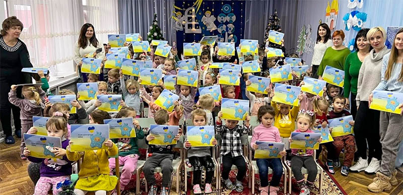 Preschool-aged children sit in chairs and hold up their new books over their heads. Adults are seen in the back and around the edge of the classroom.