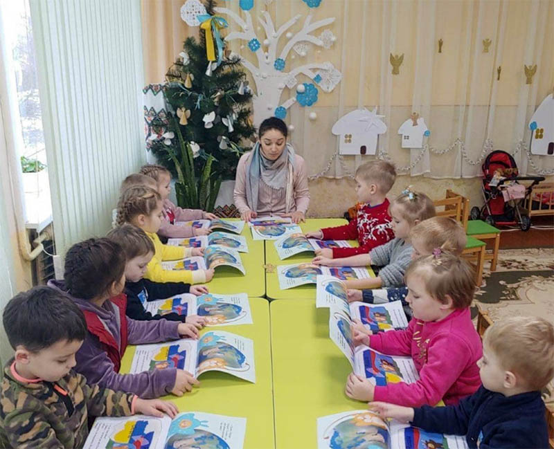 A young female teacher sits at the head of a table full of preschool aged children, helping them understand the book they all have.