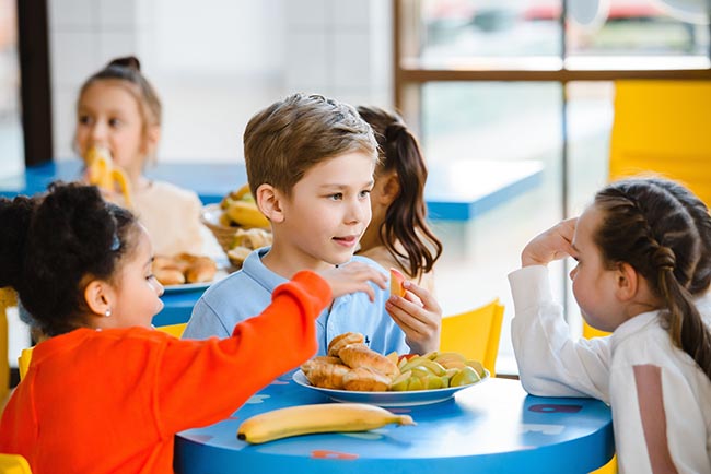 A group of kindergarten-aged children sit at blue tables eating snacks.