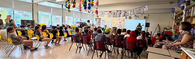 Wide angle shot of Kim Moberg singing and playing guitar up in front of a classroom of young children.