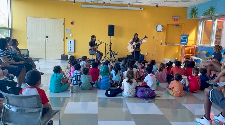 Kim Moberg with her acoustic guitar in front of a group of school children seated in an small performance room. A percussionist is seated next to Kim.