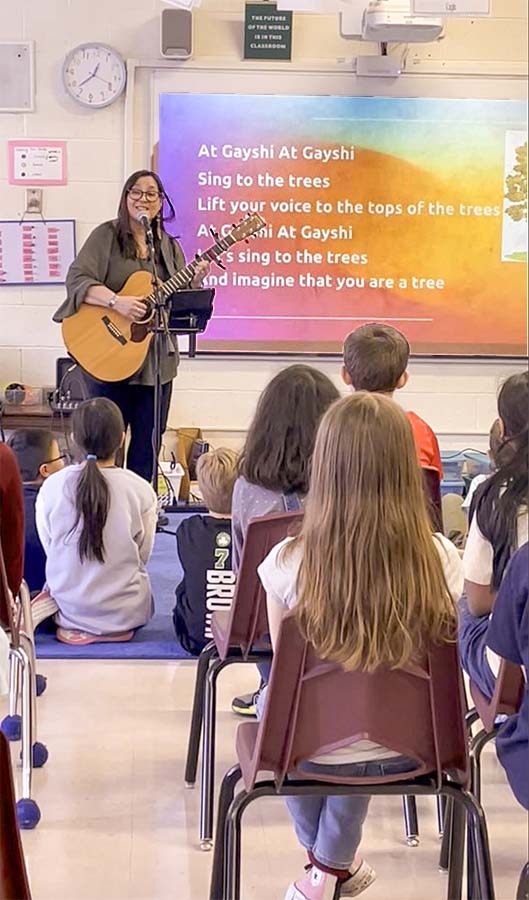 Kim Moberg performing with her acoustic guitar in front of a school room of children seated at their desks. A brightly colored slide is on the screen behind her with lyrics.