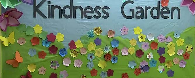 Felt banner reading Kindness Garden on a sky blue backdrop with a green expanse below with felt flowers and butterflies attached.