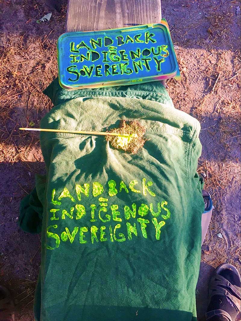 A stencil, paint brush, and stenciled t-shirt on a wood bench. The stencil reads: Land Back, Indigenous Sovereignty