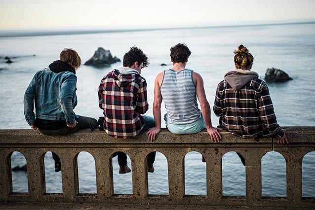 Four teen men seen from the back sit on a stone fence with their feet dangling over the ocean below.