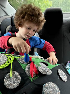 A boy in a car seat plays with his "manhole cover" board game he's created of felt and yarn.