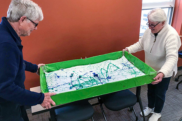 2 seniors hold a box between them with a marble-rolling painting inside
