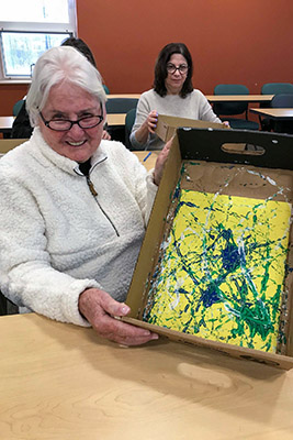 A senior woman holds up a small box with her marble-rolling painting inside