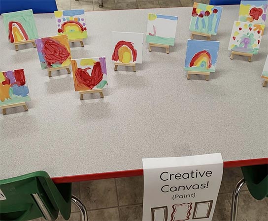 A classroom table with tiny easels with small, square watercolor paintings set on each one.
