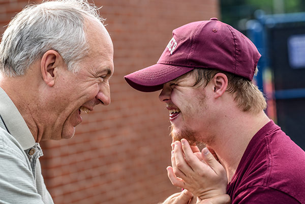 A grandfather stands face-to-face with his disabled grandson. Both are laughing. The son wears a burgundy shirt and baseball cap.