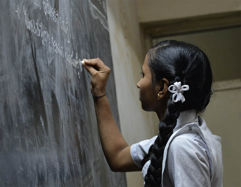 A brown-skinned girl with pigtails stands at a blackboard writing with chalk