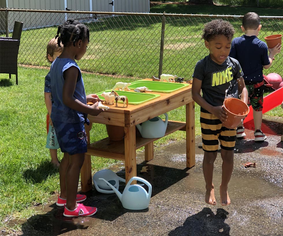 A group of kindergarten children play outside at a mud kitchen with buckets and watering cans.