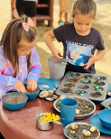 A young girl and boy create mud muffins and a cake in the outdoor mud kitchen