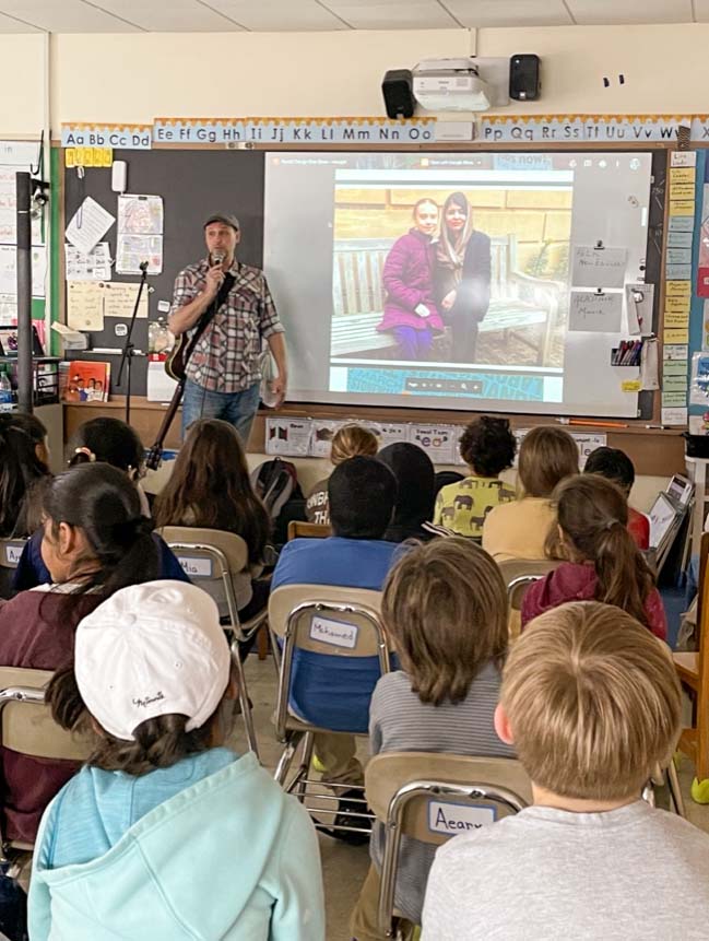 A man with a guitar stands in front of a class of elementary school children speaking into a mic, discussing the slide show on the video screen next to him.