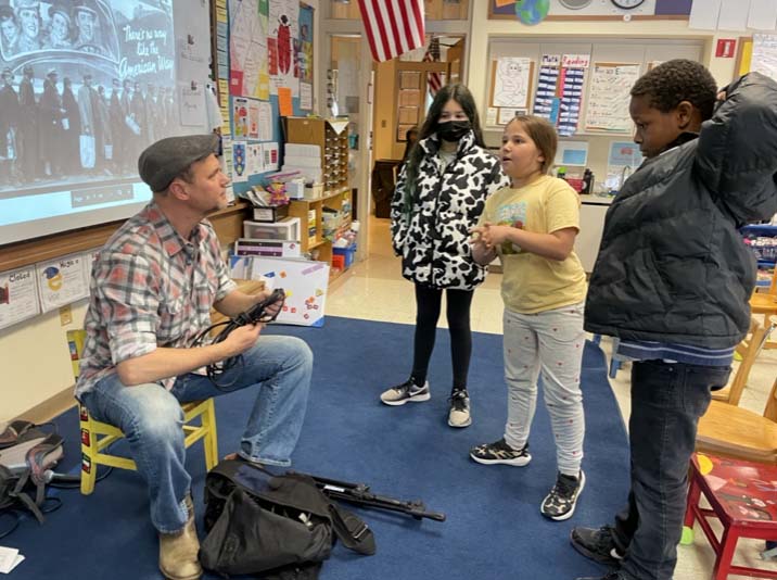 A man sits speaking to 3 fifth-grade students about music and social change in a classroom.