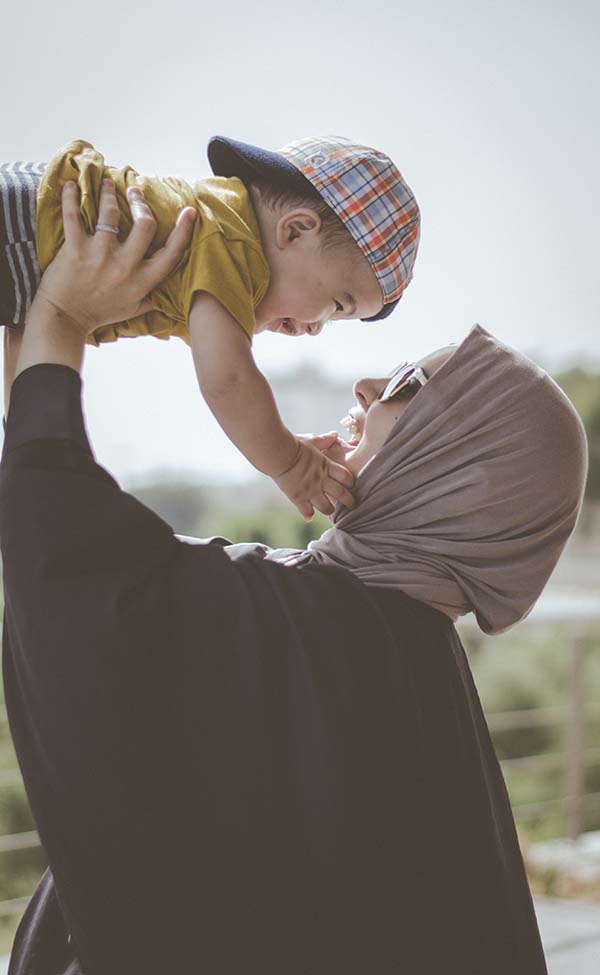 A Muslim mom swings a little boy up over her head. She wears a head scarf; he has on an infant baseball cap, placed backwards