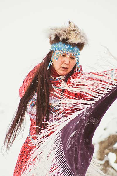 A Native American woman in full headdress and gown performs a native ceremonial dance.