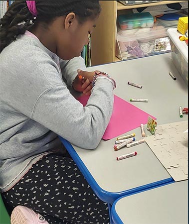 A school-aged girl uses colored pastel sticks on pink construction paper.