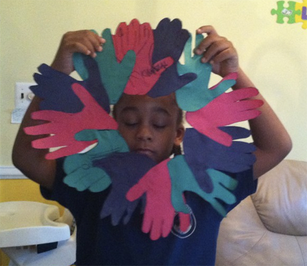 A black bow holds up a wreath to his face made out of hand shapes cut out of colored construction paper.