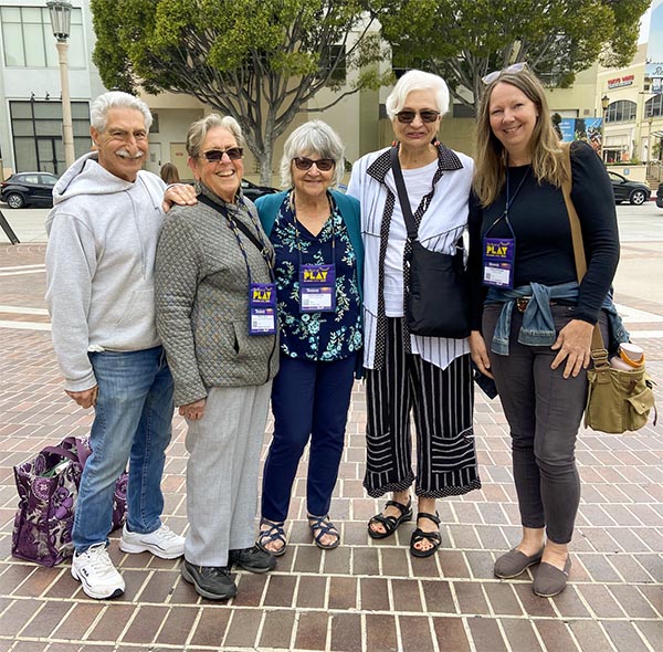 5 middle-aged people stand arm-in-arm smiling at the camera. They are standing on a brick area with an intersection behind them.