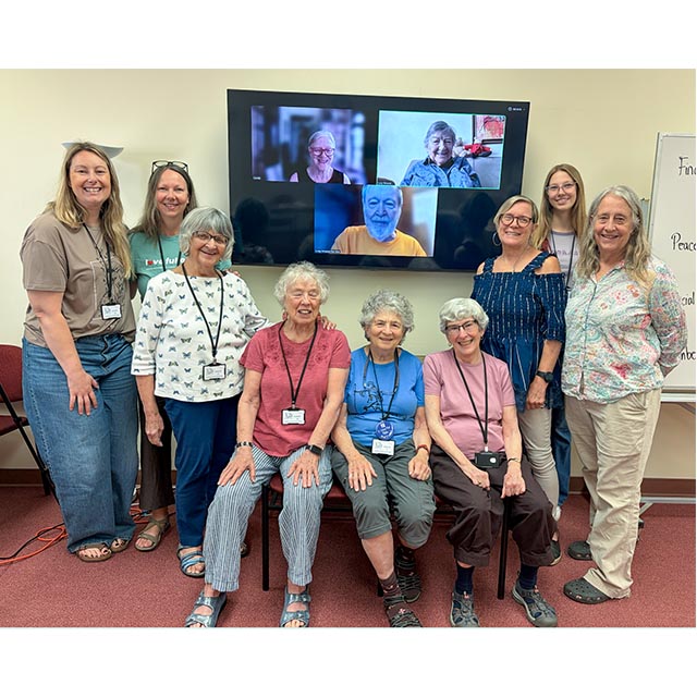 A group photo of the PEACE board members at their annual retreat. Nine members stand in front of a large, wall-mounted screen with three members who joined remotely.
