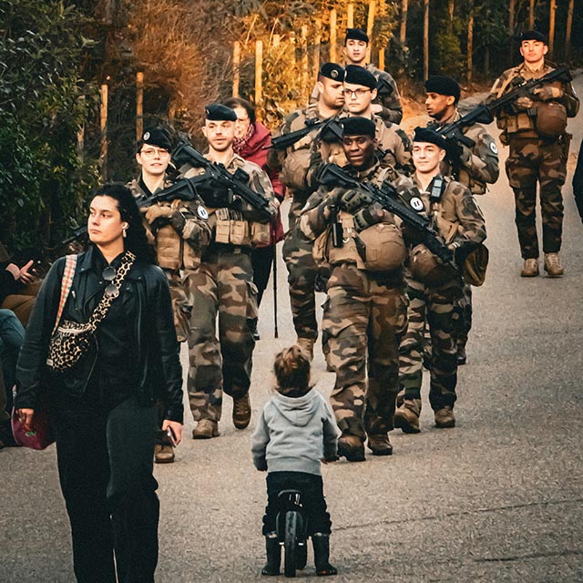 A small child on a bike looks up as soldiers march past in the street.