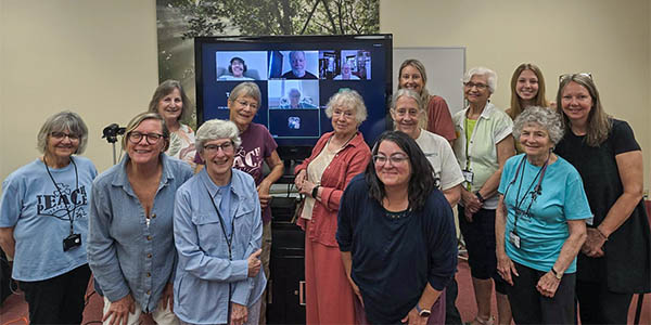 A group of middle-aged adults standing in a classroom with a large screen behind them showing the Zoom squares of other meeting attendees.