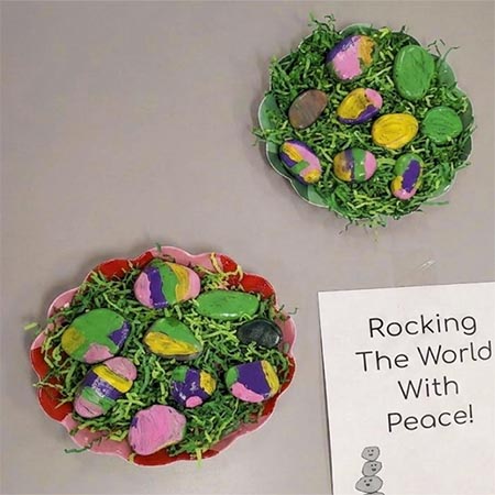 Rocks painted to look like Easter eggs sit in bowls with crinkled green paper. A sign next to the bowls reads, "Rocking the world with peace."