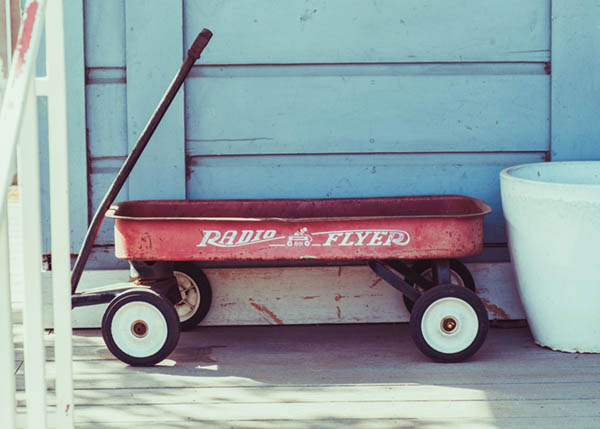 A red metal Radio Flyer wagon sits on a wood porch in front of a light blue wall.
