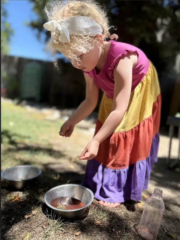 Soft-focus image of a young girl wearing a colorful dress. She leans over a bowl of water, carefully placing leaves in the bowl.